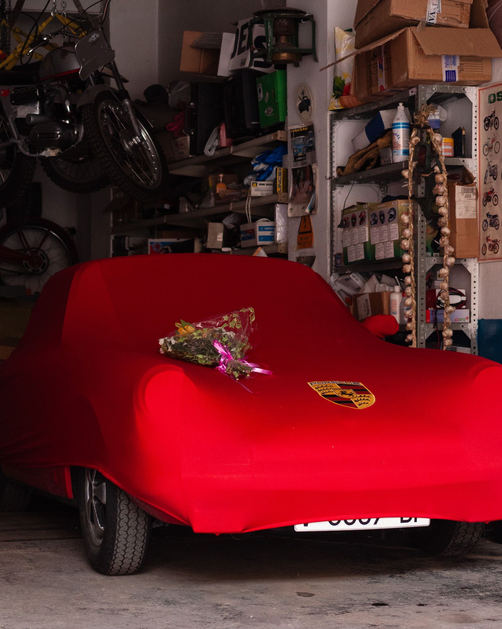 Porsche covered with a red cover and flowers on the bonnet in a garage with classic motorcycles