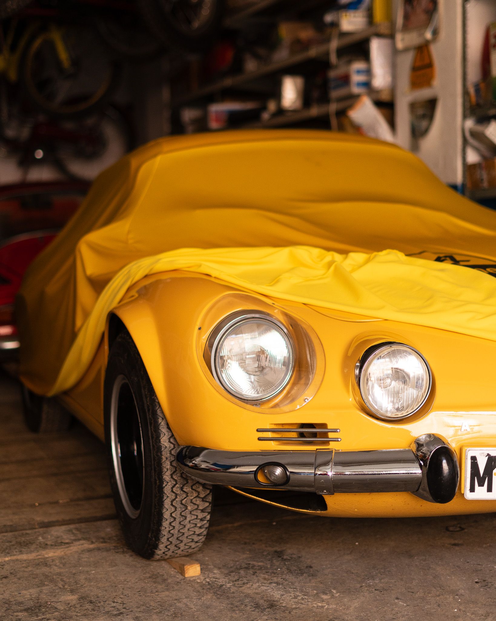 Front of an Alpine A110 peeking out from under a yellow cover in a garage full of motorcycles